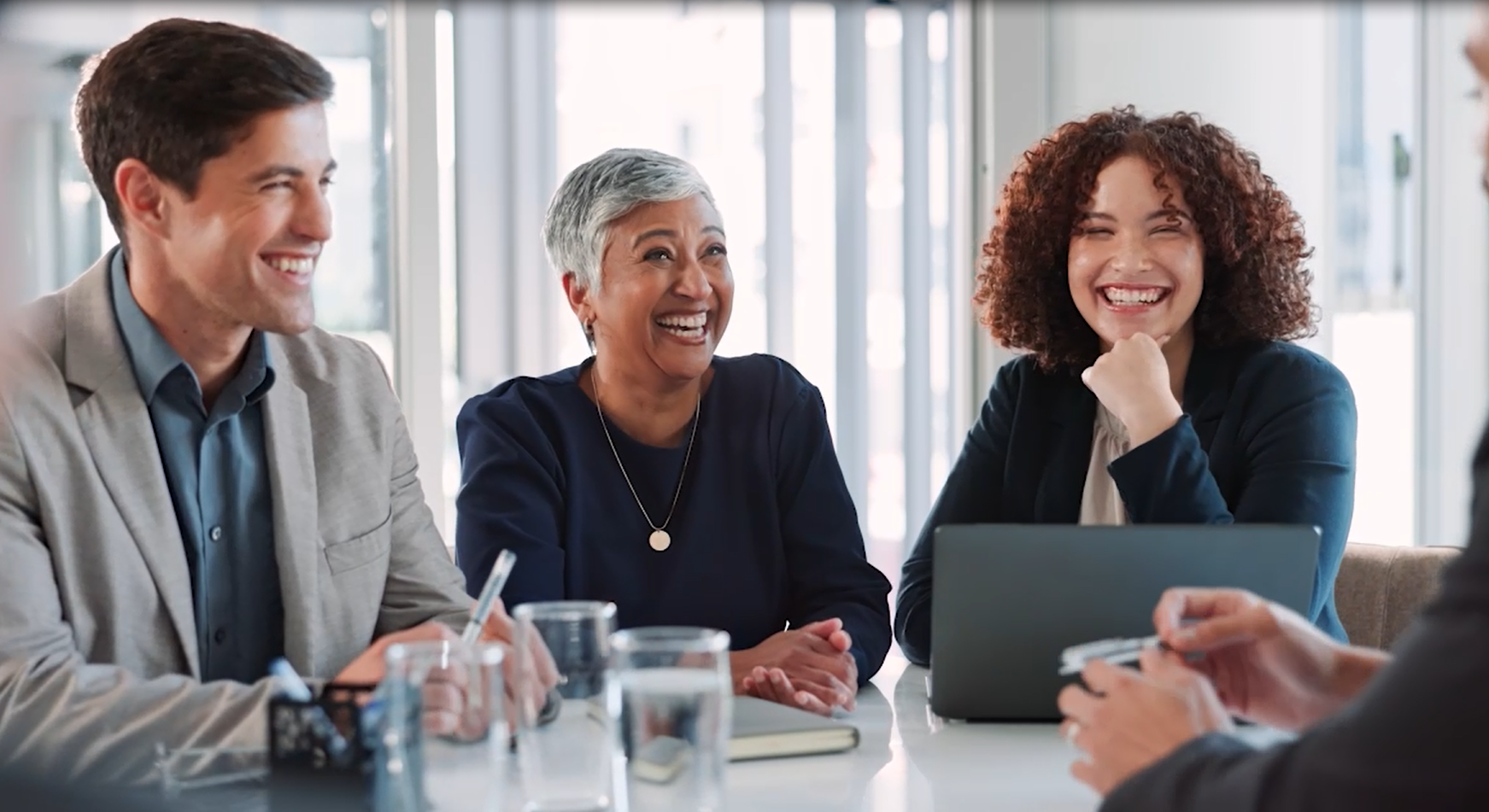 Ein Team bestehend aus drei Leuten unterhält sich lachend an einem Meetingtisch.